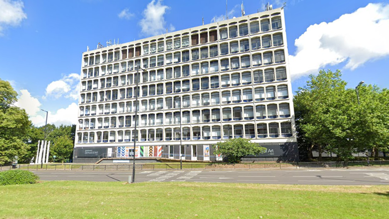 A high, brutalist building on the side of a main road. The sky is bright blue in the background and there are trees and grass around the building. There are several masts on top of the structure.