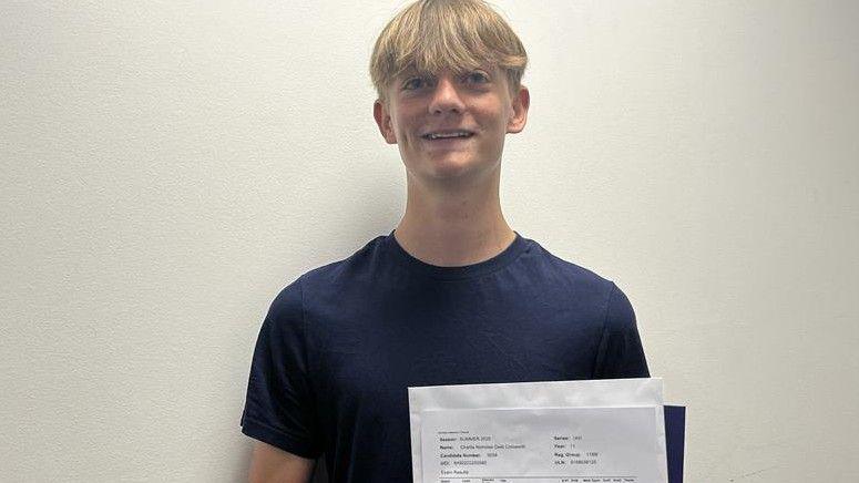 A 16 year old caucasian boy with blonde hair smiles at the camera in a navy blue t-shirt whilst holding his GCSE results paper.
