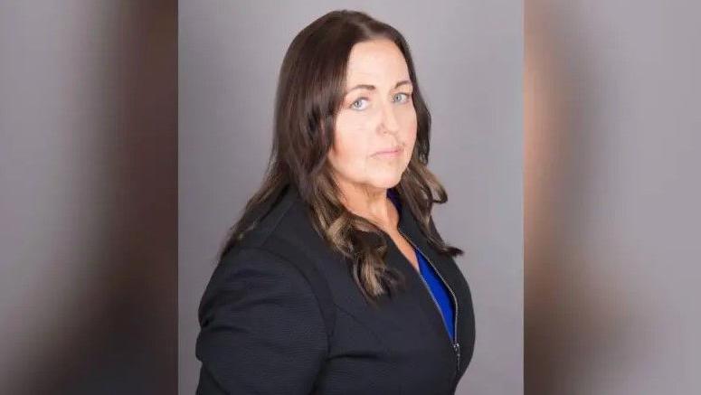 Joanne Monk, a woman standing to the side with her head facing the camera. She has shoulder length brown hair and is wearing a black suit.