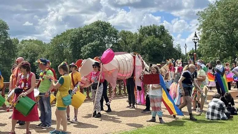 A large group of people dressed in colourful clothing marching through a park in a Pride parade. Some can be seen holding drums, flags and various displays.