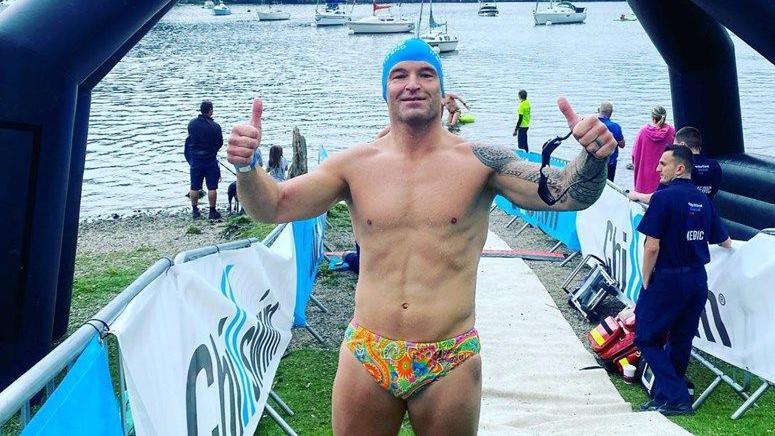 A man in multi-coloured swimming trunks sin front of a large body of water - posing with his thumbs up and wearing a blue swim cap.