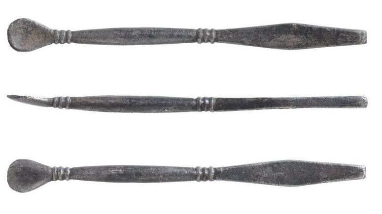 A silver stick with a spoon shape on one end and a rectangular end the other. Three round circles wrap around each end. The object is photographhed three times, the middle image shows it from a side angle.