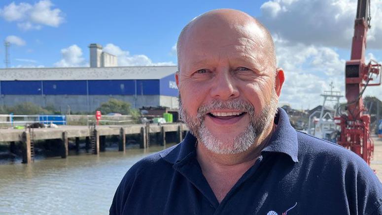 Steven Williamson is standing outside alongside the dock at King's Lynn. He has a short grey beard, is wearing a blue polo-shirt and is smiling. To the left is the Ouse estuary river and on the other side is a large warehouse.