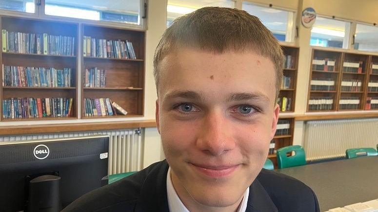 Josef has short, light brown hair and blue eyes and is wearing a black bl;azer and white shirt