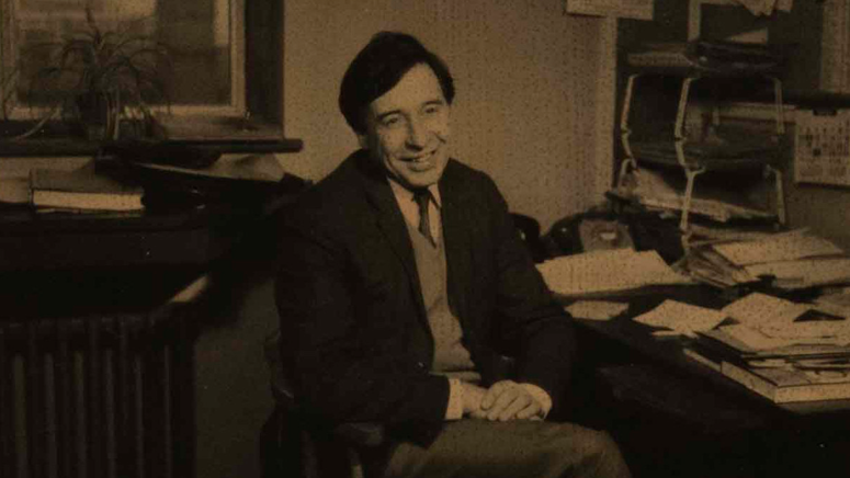 Sepia-toned photo of Prof Edwards in a suit smiling while seated in a cluttered office, with papers, calendars, and a window in the background. Taken at the University of Cambridge Physiology Department.