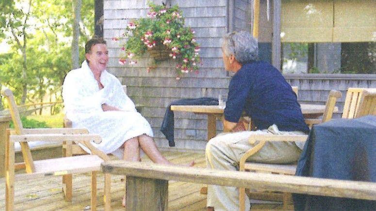 A copy of an undated picture of Peter Mandelson sitting in a white robe laughing while sitting opposite Jeffrey Epstein, who wears a blue top and cream chinos, on a wooden deck.