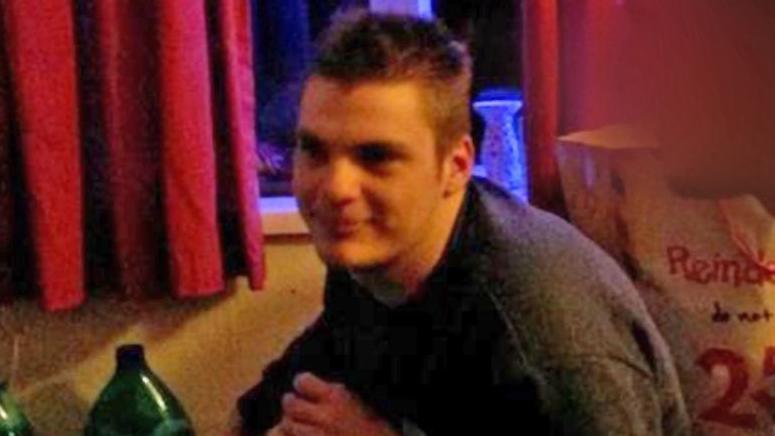 A grainy picture of Jamie Harding, who is wearing a blue sweater. He has short brown hair and is smiling. Behind him is a blue lava lamp and red curtains. He is sat in what looks like a bedroom.