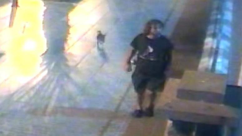 A CCTV image of James Nunan, who has long hair and is wearing a black T-shirt and shorts, walking along a wide pavement.