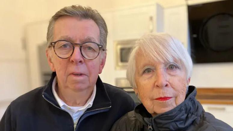 An elderly man with grey hair and glasses stood beside an elderly woman with short grey hair and red lipstick, looking into the camera. They are stood in a large white kitchen.