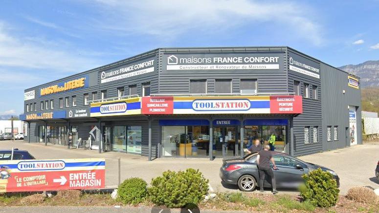 The French branch of Toolstation in a grey building built on a corner.  There are Toolstation logos above the doors with French words outlining what the shop sells. A sign in the foreground indicates the entrance to the shop.
Two men are getting out of a car in the car park.