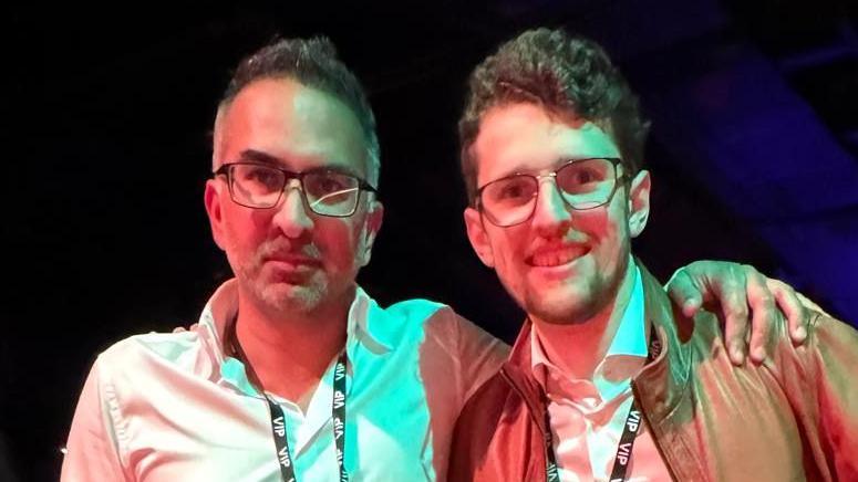 Shiv Malik and Joseph Reeve stand together and smile at the camera. Shiv has short dark hair with some grey hair and wears glasses along with a white shirt and lanyard around his neck. Joseph has short curly hair and wears glasses along with a brown leather jacket and white shirt. He also has a lanyard around his neck. Shiv rests his arm around Joseph's shoulders.