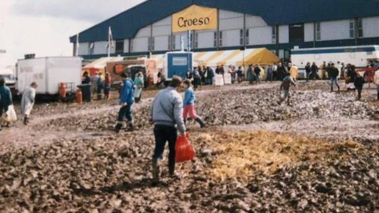 Dyn yn cerdded drwy fwd ar Faes yr Eisteddfod