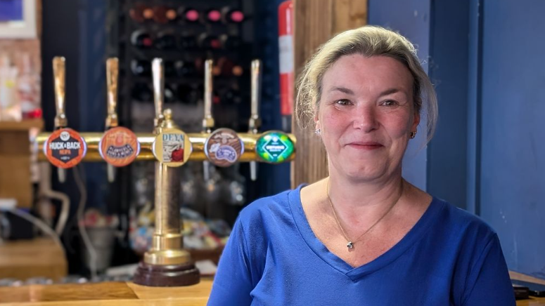 A wide shot of Fiona Hornsby standing in front of the bar at her pub. She has tied-back blonde hair and is wearing a blue top.
