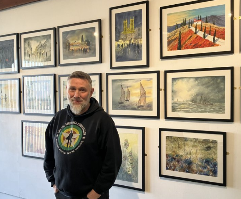 Tim has short dark hair and a grey beard. He is wearing  black hooded top with a green circular print and is standing in front of a wall on which a series of framed paintings are hanging.