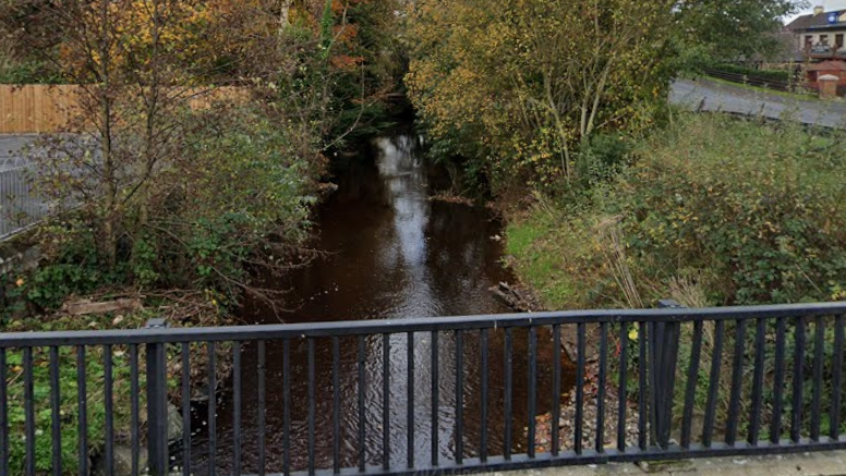 A grey bridge railing is seen overlooking a shallow river basin.
Hedges and trees can be seen either side of the river.