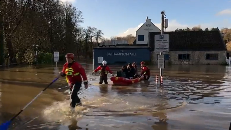 Fire crews wading through water pulling a boat with five people and two dogs inside. Bathampton Mill, surrounded by flood water is in the background