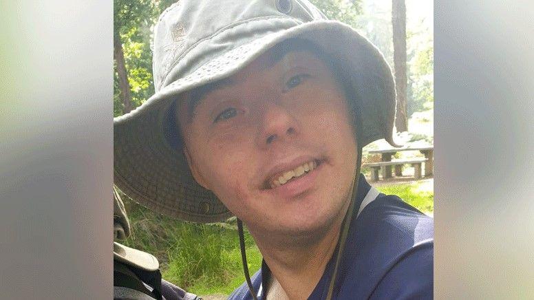 A close-up of a man's face. He is wearing a white hat and a blue shirt and is smiling at the camera. There are some trees and a wooden bench in the background.