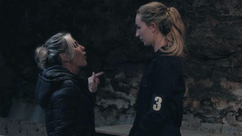 Michaella McCollum being interrogated as part of the final episode of Celebrity SAS. A small, blonde instructor wearing a black puffed coat points her finger at McCollum in a dark, stone wall room. McCollum stands on the right looking down. She is wearing a dark jacket with 3 on an armband. She has her long blonde hair tied back.