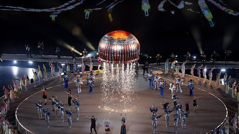 A giant teacake, wrapped in red and silver packaging, rises into the sky with sparks firing from underneath it. Various dancers and performers stand beneath it on the floor, performing a routine.