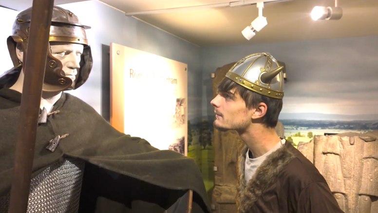 A profile view of a young man wearing a Viking helmet and clothing. He gazes at a mannequin holding a long sword, similarly dressed.