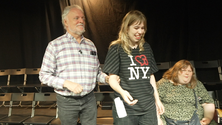 Ian Hamilton, who is blind and has grey hair and a beard, is wearing a checked shirt and grey jeans. He is holding the arm of a woman in a black T-shirt as he is led around a stage in a theatre