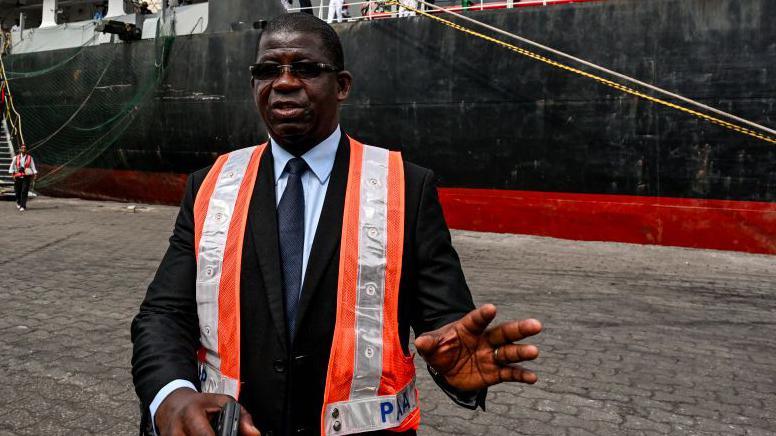 Abidjan port boss Hien Yacouba Sie speaks to reporters. He is wearing a hi-viz tabard. Behind him in the outer harbour is the Zimrida vessel.