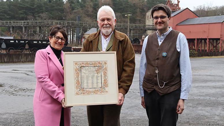 Two men and one woman, against a background of an old pit building.
The man in the middle, who has grey hair and a beard and is wearing a brown jacket is holding a frame containing a serviette marked with a list of names and a floral border. To his left is a woman with dark short hair and glasses wearing a pink coat. The man on the right is dressed in a light blue shirt and a brown waistcoat with a watch chain - in the style of a miner from the 1900s.
