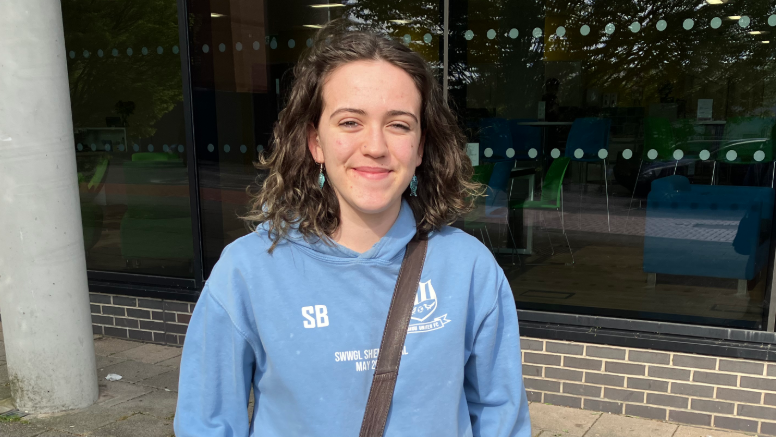 Merch mewn hoodie glas golau a bac dros ei hysgwydd yn sefyll tu allan i adeilad