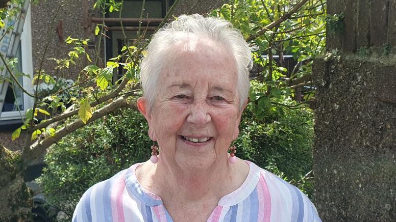 A lady with short grey hair smiles at the camera as she wears a pink, white and purple striped shirt, and pink dangly earrings. Behind her is a green bush and tree with green leaves.