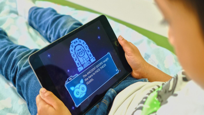 An over-the-shoulder photo of a young boy sat on a bed with blue cloud bedding. He is wearing jeans and a t-shirt. He is holding a black IPad and is playing a game on the Lumi Nova app. 