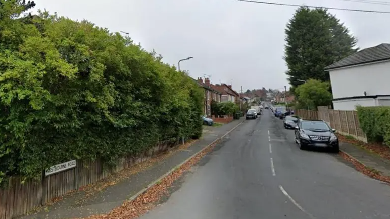 There is a hedge on one side and houses on both sides further down the street with cars parked on both side of the road.