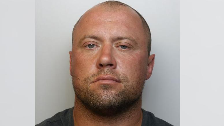 A mugshot of Ashley Harris. He is wearing a black t-shirt and standing against a grey background. 