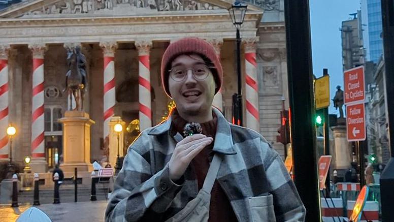 Tom wearing a red beanie hat, glasses and a checkered shirt while speaking into a microphone.