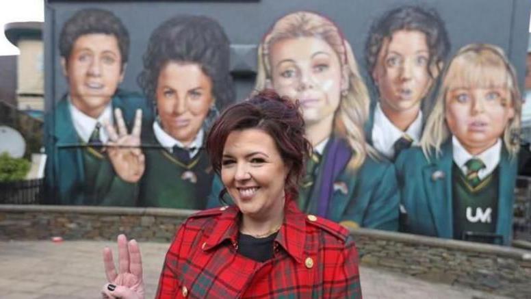 Derry Girls creator Lisa McGee, in red and green check jacket and holding up three fingers on her right hand, stands in front of the Derry Girls mural. The mural depicts the characters from her hit comedy show.
