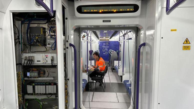 A technician works at a desk in an empty carriage, wearing an orange high-visibility vest.