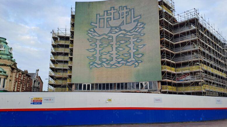 A construction wall along the front of a high building covered in scaffolding. A mosaic mural depicting three white ships and waves outlined in blue covers most of the front of the multi-storey building. 