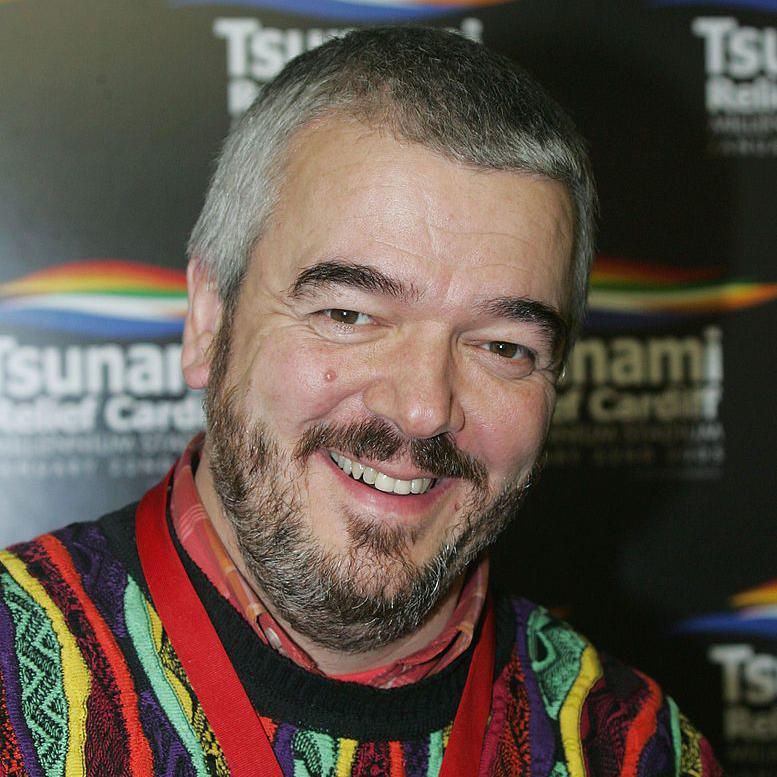 Paul Sergeant with grey hair and a black beard and a colourful jumper smiles for the camera at the 2005 concert