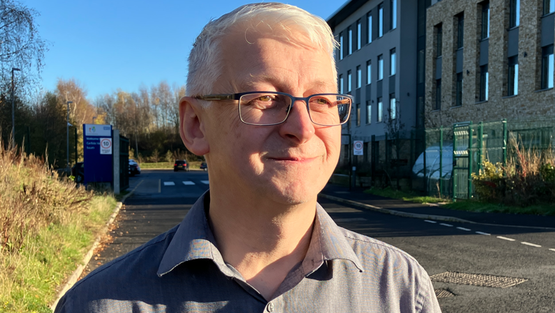 Dr Robert Westgate, with short white hair, glasses, and wearing a grey shirt, looks to the right of the camera, as he stands on a road outside a large building on the right, with rectangular windows. There is a car park entrance behind him. 