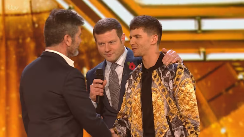 Leon Mallettt on The X Factor stage wearing a golden jacket and shaking hands with Simon Cowell. Dermot O'Leary has his arm around Leon's shoulder.