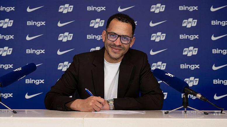 Photo showing a smiling Liam Rosenior signing his Chelsea contract, with advertising logos behind him on a wall also bearing the words Chelsea Football Club and two club badges