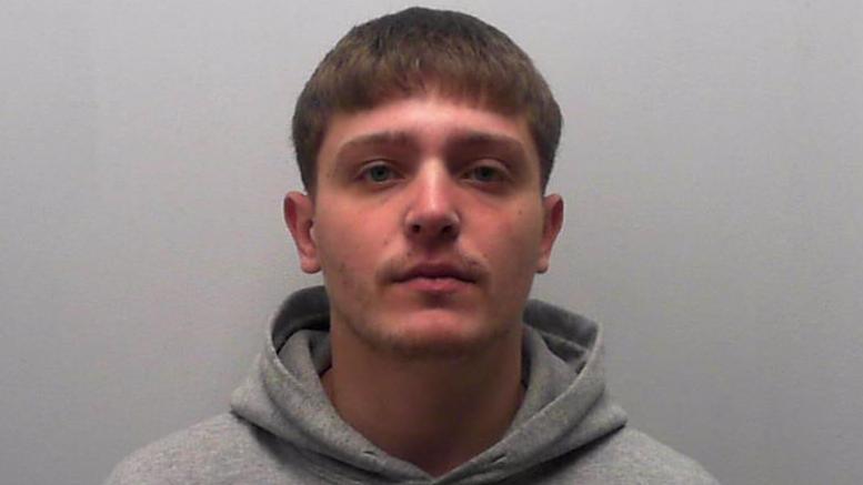 Image of a young man in grey hoodie at a police station. He has short brown hair. He is standing in front of a grey background.