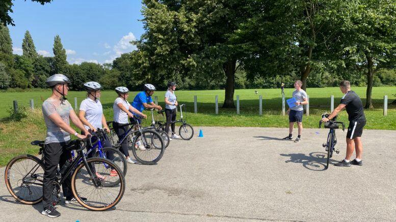 Several people on bikes looking towards a man holding a clipboard. They each wear a safety helmet. There are trees in the background.