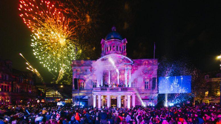 A large crowd watches red and yellow fireworks light up the night sky over Hull City Hall, a grand Edwardian building with columns, a balcony and a dome. The building is lit up in purple by light projections.