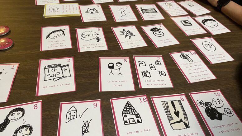 Several cards laid out on a wooden table. They are all numbered and have children's drawings on them and writing including faces, a house and objects.