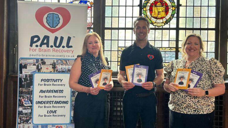Three people holding a purple and a yellow leaflet stood in front of a stained glass window inside a building. They are stood next to a banner with multiple images and reads 'P.A.U.L For Brain Recovery'. The woman on the left is wearing a black spotty blouse and black trousers. She has long blonde hair. The man is wearing a black polo shirt with a red and blue logo on and black trousers. He has short brown hair. The woman on the right is wearing a black and white patterned blouse and black trousers. She has light-coloured hair tied back. They are all smiling at the camera.