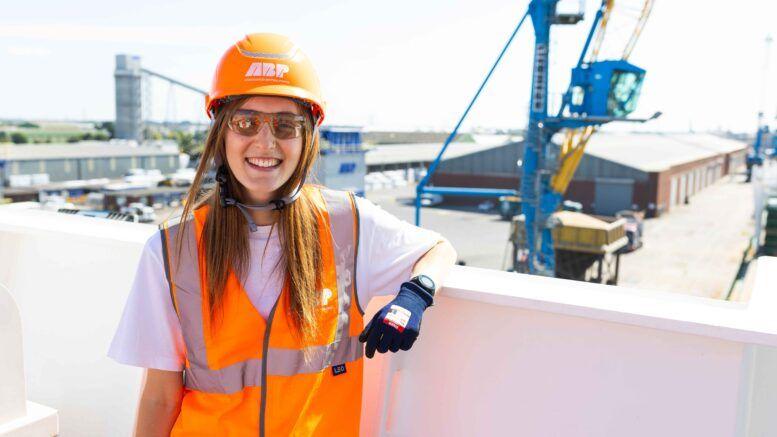 A smiling Carrie Grimbleby leaning on a wall at a port wearing an orange hi-vis jacket and hard hat with the ABP logo on it