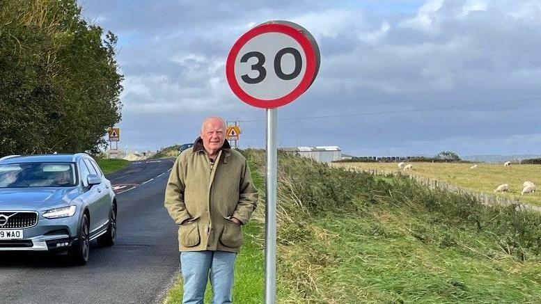 John Rhind is standing next to a 30mph zone sign on a rural road as a car speeds past