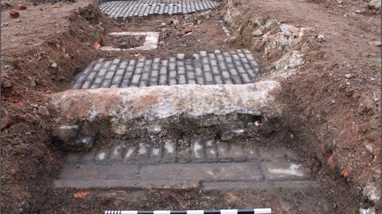 An area of excavated ground with grey stone setts at the lowest level. There is a black and white measuring tool in the foreground.