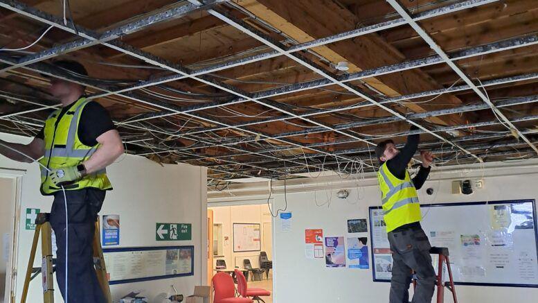 Two men standing on step ladders at opposite sides of a room, both wearing high-vis vests over black tops and dark grey trousers. They are attaching wires to the beams of a bare ceiling in a community centre room. There is a long whiteboard on the far right wall and a green fire exit sign on the left wall.