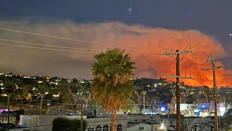 A view of part of LA where homes and buildings can be seen. In the distance a large plume of smoke billows into the sky and it has an orange glow within it from the wildfires below.